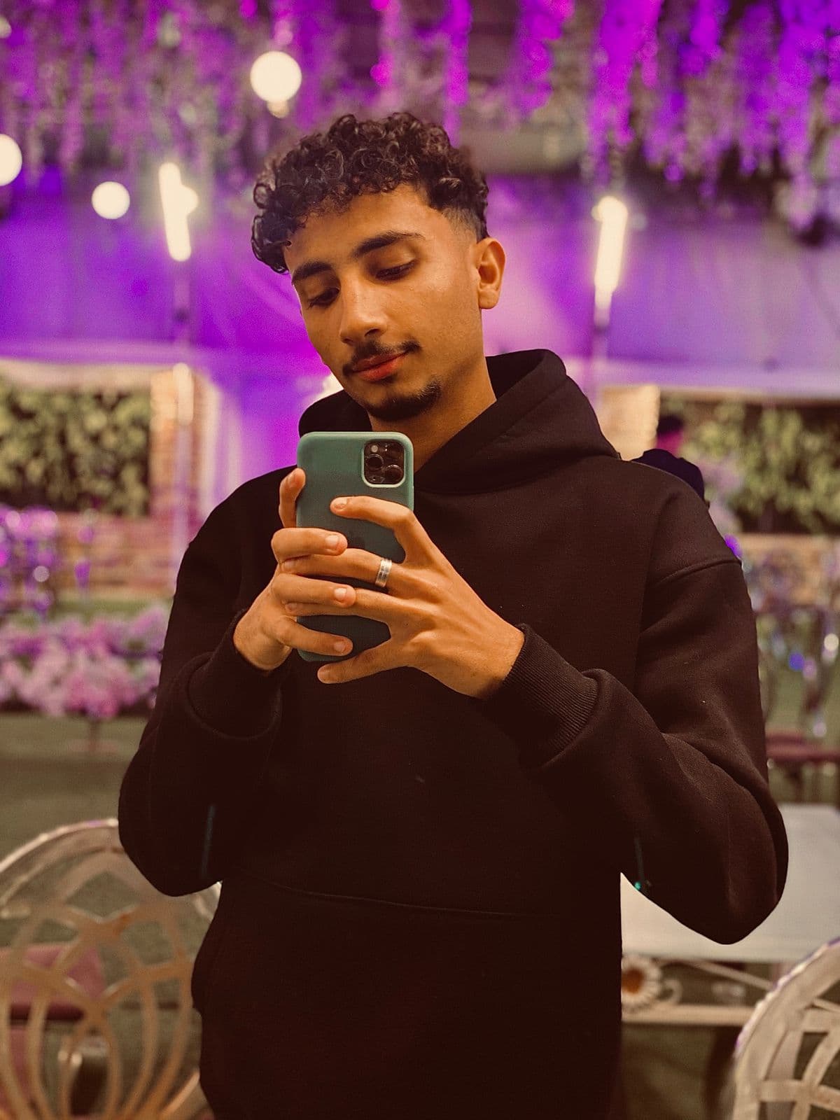 Homme aux cheveux bouclés prenant un selfie en sweat noir devant un décor festif violet.