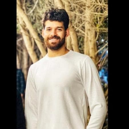 Homme souriant aux cheveux bouclés et barbe, portant un chandail blanc devant des arbres.